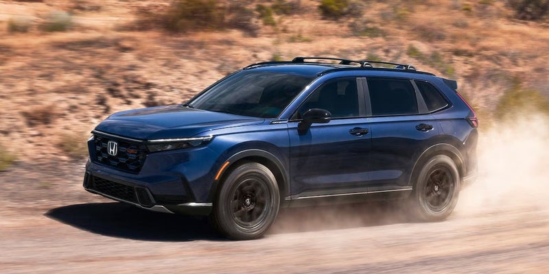 Dark blue SUV with a roof rack driving right to left on a dirt road, kicking up a large cloud of dust.