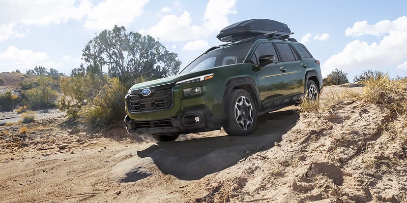 A dark green Subaru Forester with a roof cargo box drives on a sandy, rocky off-road trail.