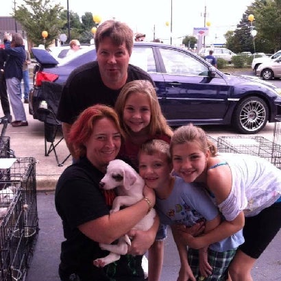 A family poses with a white puppy, smiling. A dad and three kids stand behind a kneeling woman holding the puppy.