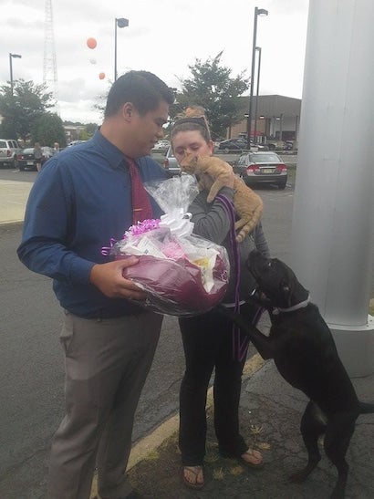 A man holds a large gift basket next to a woman holding an orange cat, while a black dog jumps up at her.