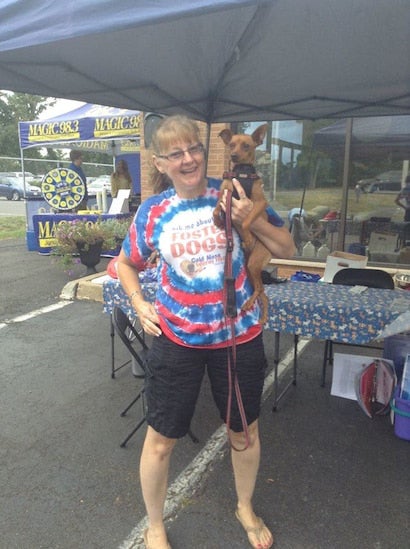 A smiling woman in a Fostered Dogs tie-dye shirt holds a small brown dog with a harness under a tent.