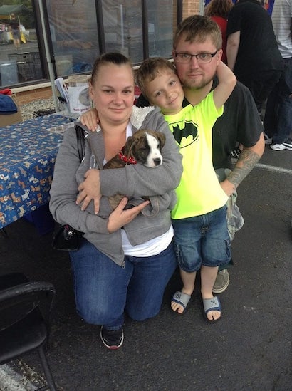 A family of three (woman, man, young boy) poses with a brindle and white puppy held by the woman.