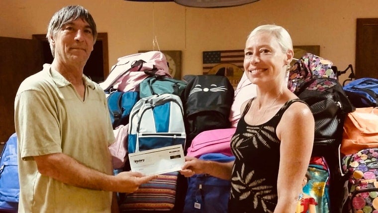 A man and woman smiling while exchanging a check, standing in front of a large pile of colorful backpacks.