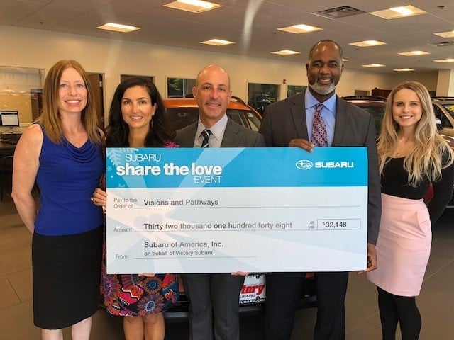 Five people in a car dealership hold a giant check for $32,148 from Subaru to Visions and Pathways.