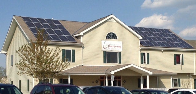 A two-story beige building with dark green shutters on windows and solar panels on both sections of the roof.