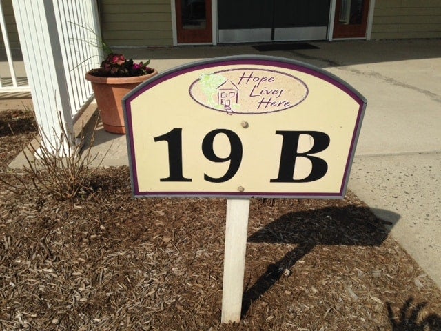 A ground sign with 19 B and Hope Lives Here in front of a building with a white railing and potted plant.
