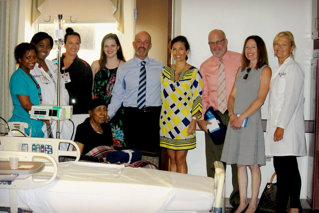 A diverse group of staff and visitors standing around a seated elderly patient in a hospital room.