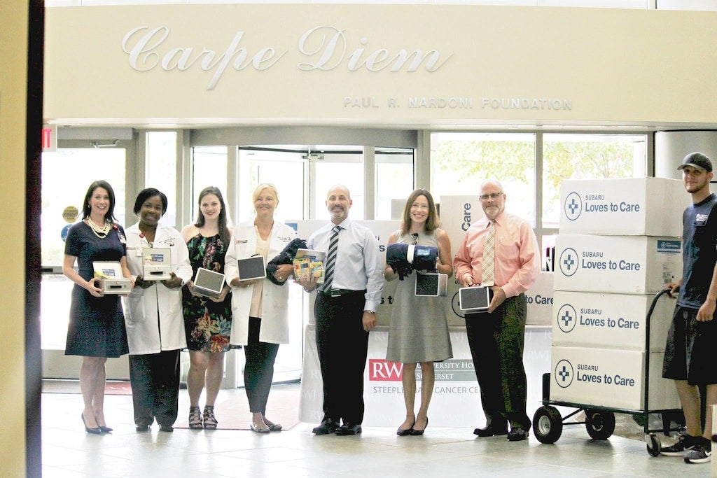 Seven adults and a young man in a hospital hall, some holding tablets, with Subaru donation boxes on a dolly.