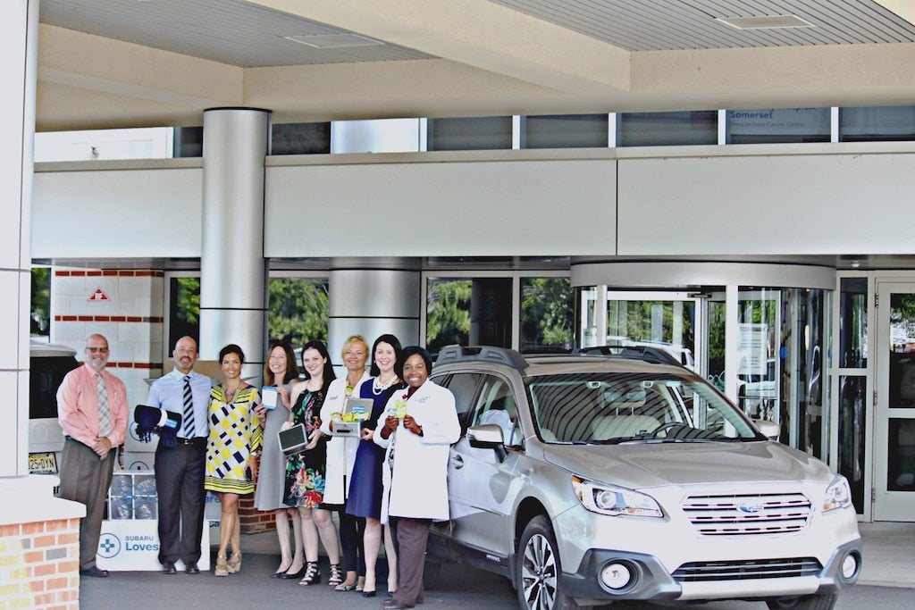 Group of eight professionals, including nurses and hospital administrators, posing with a silver Subaru Outback outside a hospital.
