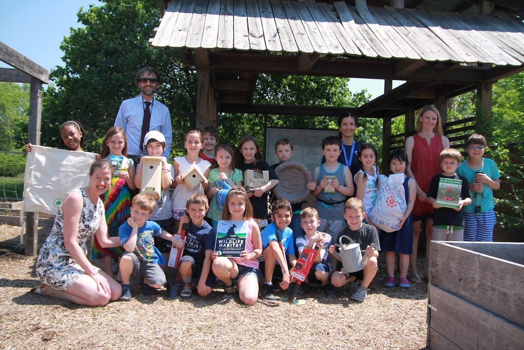 A group of children and adults pose outdoors, holding birdhouses, books, and gardening tools.