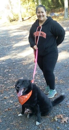 Woman in a black hoodie and black dog with an orange bandana on an outdoor path.