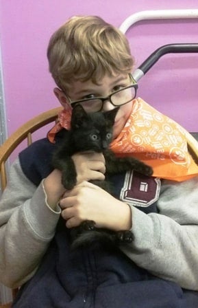 Young boy in glasses and an orange bandana holding a small black kitten.