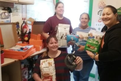 Four smiling women holding bags of dog food and treats, with open boxes on a table in the background.
