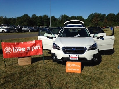 White Subaru Outback with all doors and trunk open, next to a red Subaru Love a Pet banner on a grassy field.
