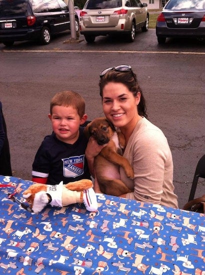 A smiling woman holds a brown puppy while a young boy in a New York Rangers shirt sits beside her.