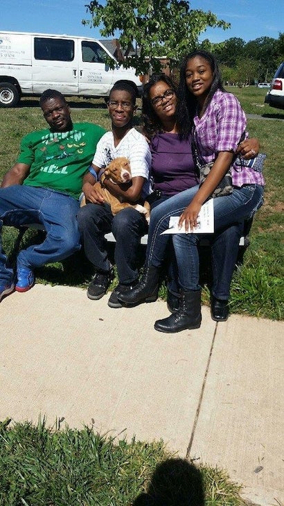 Four Black people, two men and two women, and a light brown puppy, sit on a white bench outdoors.