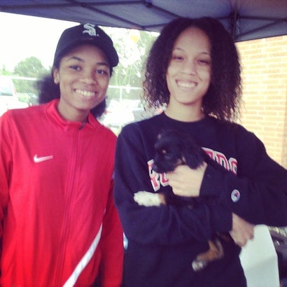 Two young women smiling, one wearing a red jacket and hat, the other holding a small black and tan puppy.
