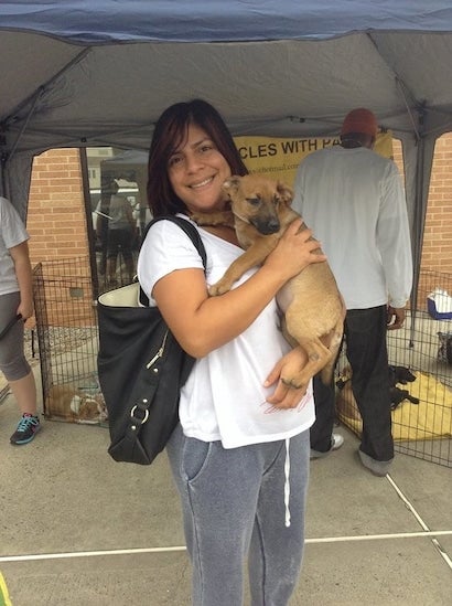 A smiling woman with dark hair holds a light-brown puppy at an outdoor rescue event with dog crates in the background.
